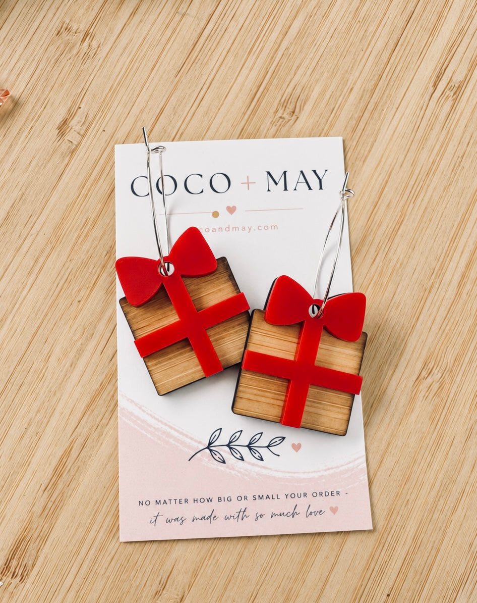Christmas present earrings featuring golden gift boxes with bright red bows hanging from gold-tone hoops, displayed on a Coco & May earring card