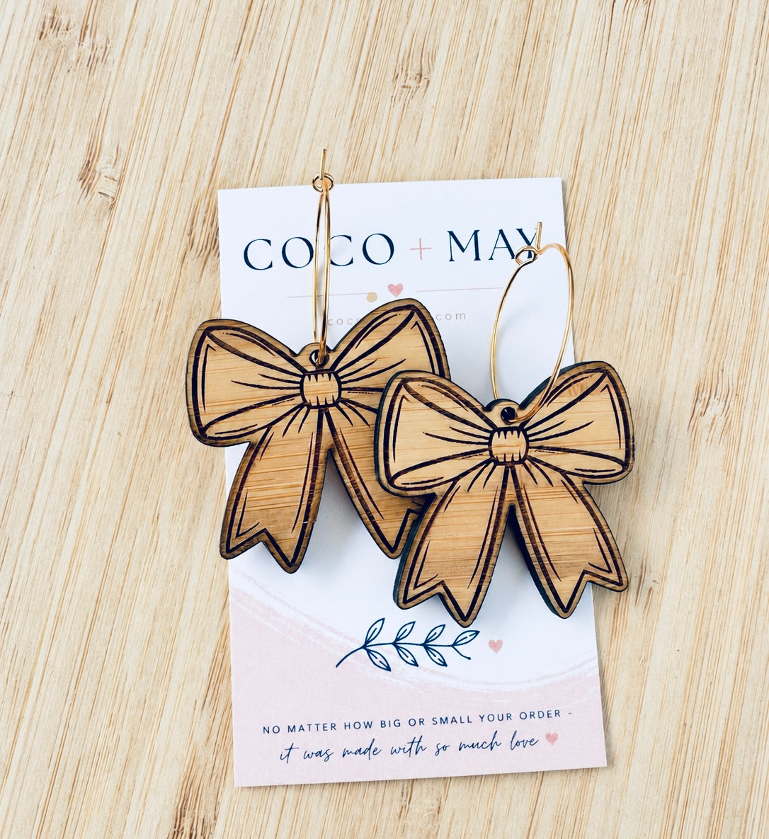 Natural bamboo bow-shaped dangle earrings with engraved detailing and dark outlines, displayed on a Coco & May jewellery card