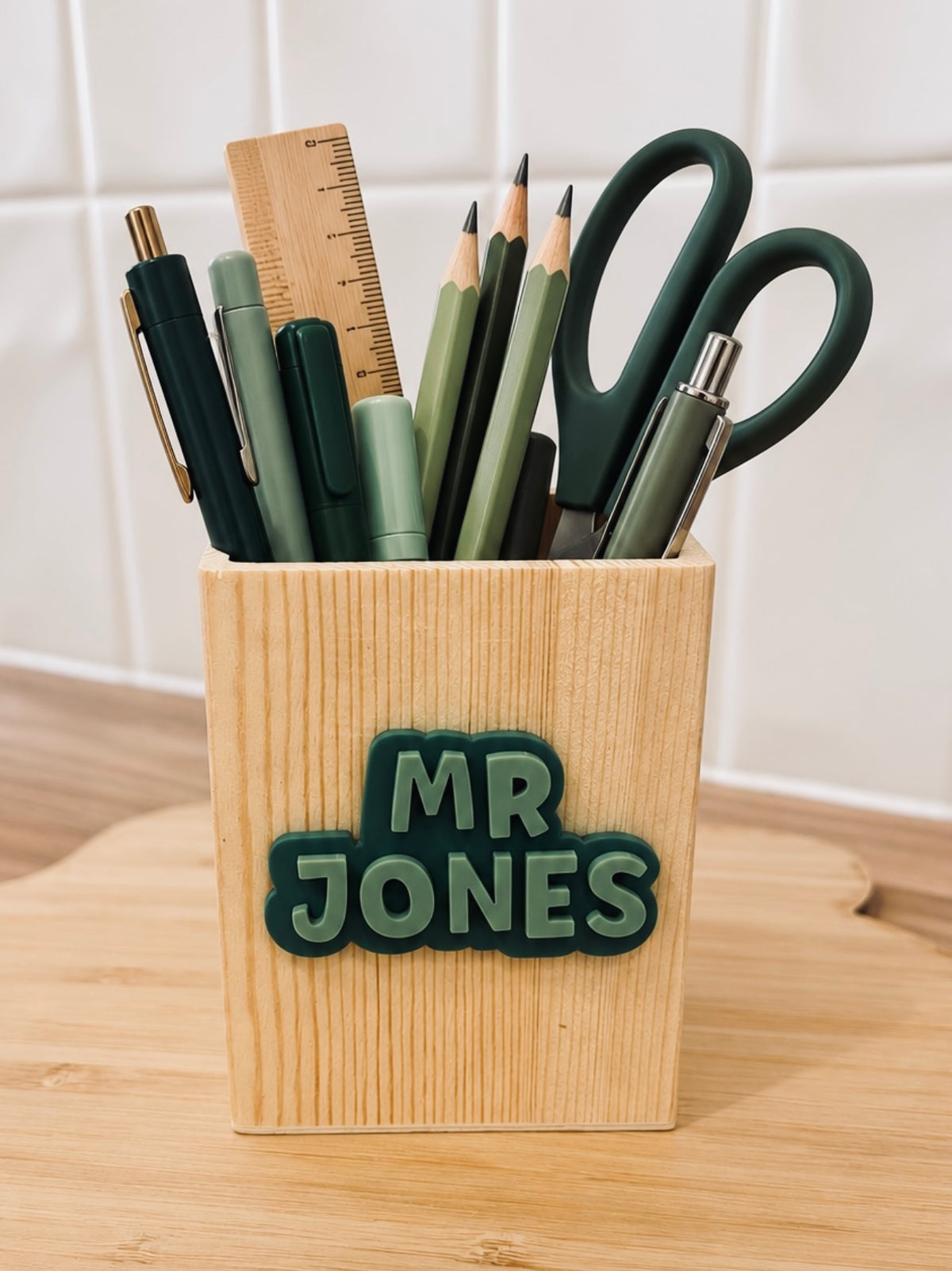 Personalised wooden teacher pen holder with raised dark green Mr Jones lettering, displayed with pens, pencils and scissors by Coco & May